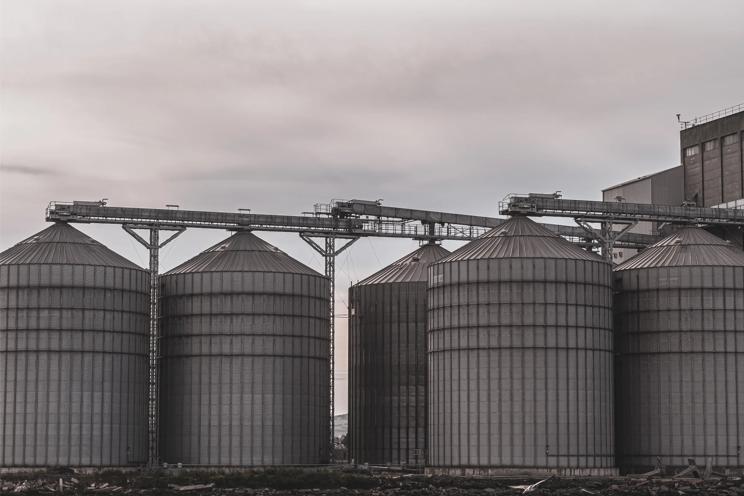 Steel silo bins with conveyor gantries in a port, under an overcast sky